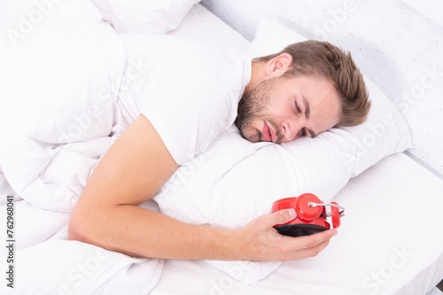 Sleepy man looking at alarm clock in hand while lying in bed. Morning awakening. Sleepy man turning off alarm clock. Waking up early. Time to wakeup and get up. Morning wakeup