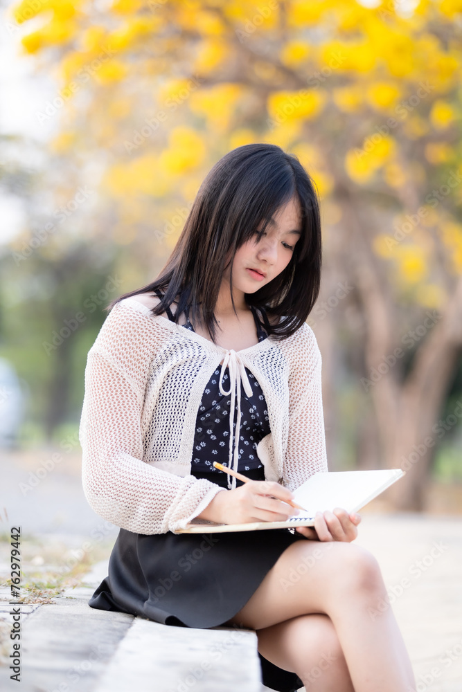 Portrait cute smiles Asian of attractive young teenage girl a holding diary book writing note at Yellow Tabebuia Chrysotricha flowers with the park in spring day at Evening background in Thailand. Portrait cute smiles Asian of attractive young teenage girl a holding diary book writing note at Yellow Tabebuia Chrysotricha flowers with the park in spring day at Evening background in Thailand.