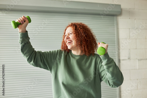 Side view portrait of ginger head having fun lifting green dumbbells, training at gym, laughing doing workout in sports sweatshirt, feeling as champion after finally finishing exercises