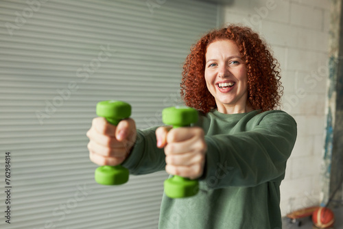 Selective focus on red head female with curls outstretching hands with green dumbbells forward, feeling happy doing workout exercise to keep her body in good shape , looking at camera