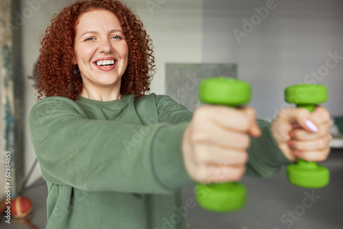Indoor portrait of selective focus on happy cheerful female face doing exercise with green dumbbells, keeping hands in front of her, training at gym in sportswear. Healthy sporty lifestyle