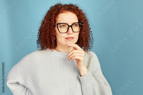 Pensive female with curly red hair in big glasses and gray sweatshirt standing against blue studio background with pensive thoughtful expression, touching chin, pondering upon idea or new project