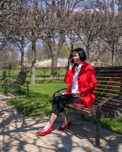 A stylish business woman sits on a park bench and talks on the phone. Makes notes in a notebook. The lady is dressed in a red cloak, white blouse, red shoes photo