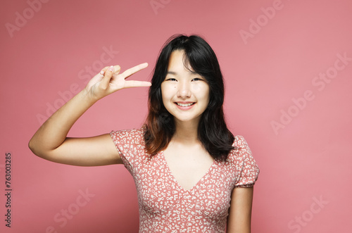 Young asian woman show victory sign , experiencing exciting emotions on pink background