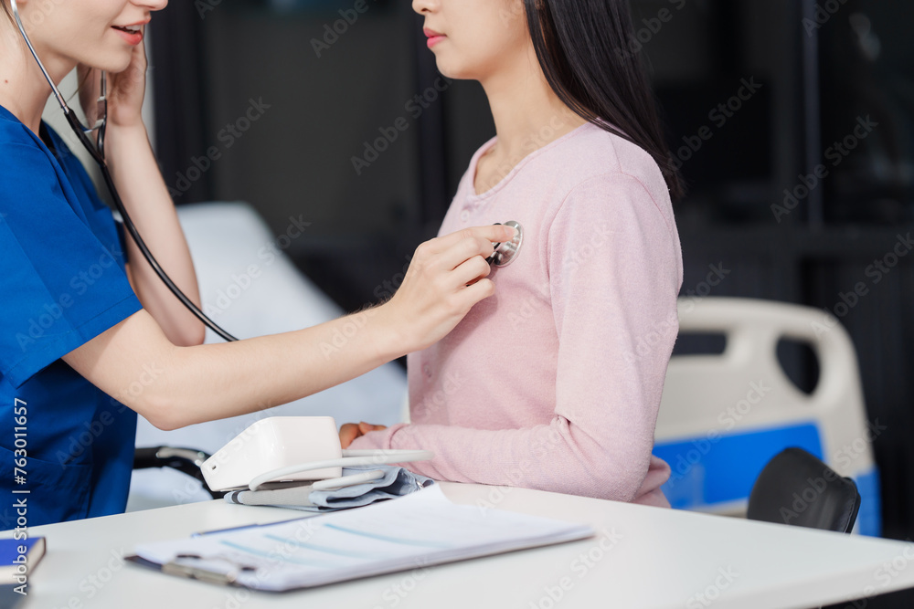 Caucasian female nurse uses a stethoscope on an Asian patient to listen to his heart rate at desk in medical office. Caucasian female nurse uses a stethoscope on an Asian patient to listen to his heart rate at desk in medical office.