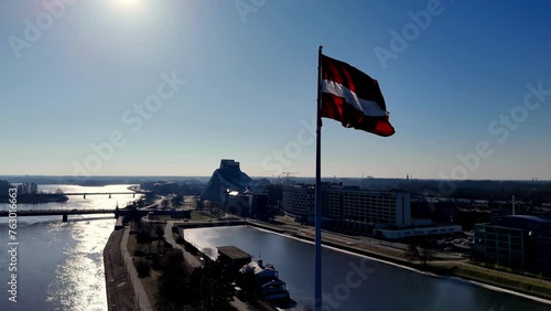 The Latvian flag on the Ganību dam, fluttering over the Daugava river, delights the residents of Riga city center. A red and white flag is flying on a pole in front of a city