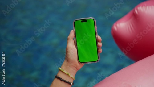 POV male hand holding smartphone with empty green screen chroma key relaxing near pool with pink flamingo background 