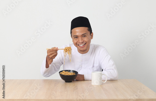 Portrait of attractive Asian muslim man eating tasteful instant noodles with chopsticks served on bowl. Iftar and pre dawn meal concept. Isolated image on white background photo