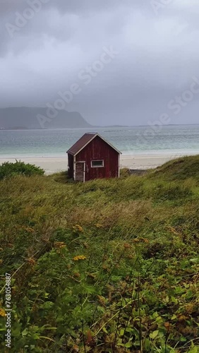 Red hut in the foreground next to a path leading to the Ramberg beach, Lofoten, Norway, during a cloudy day next to a mountains on the horizon. photo