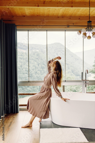 a charming girl in a brown dress is sitting on the bathroom and looking out the window. Sexy brunette is relaxing in a room with a view of the mountains,forest