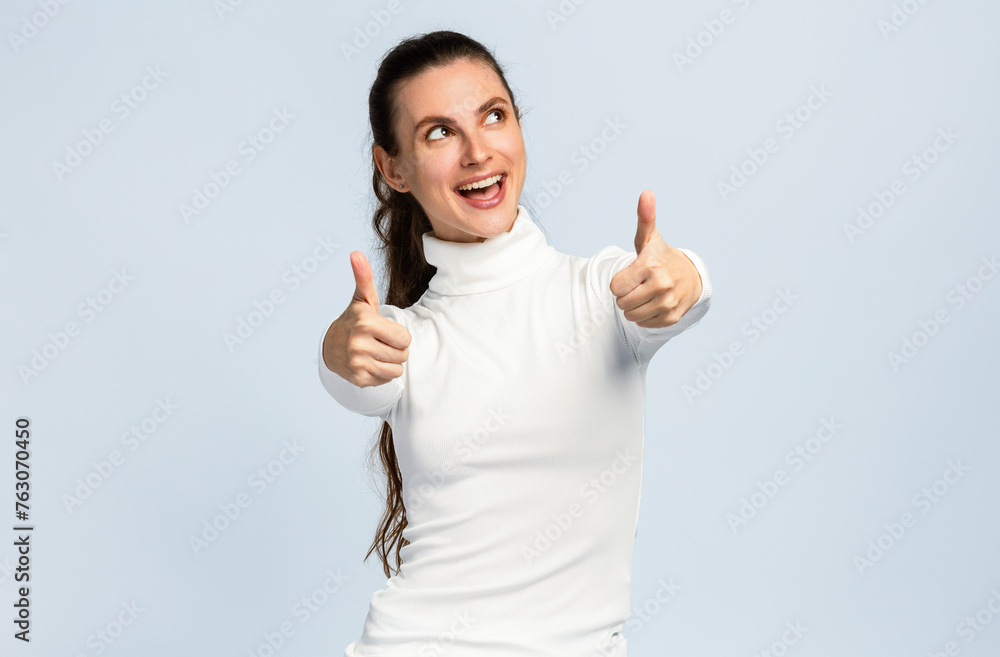 Joyful young woman make positive sign with her thumb up, positive mood, successful gesture, on light grey studio background Joyful young woman make positive sign with her thumb up, positive mood, successful gesture, on light grey studio background