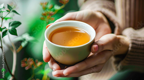 Close up of hands holding a cup with green tea, herbal medicine for colds and overall health, a warm indoor setting with plants and natural light, peaceful atmosphere