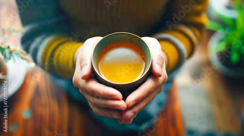 A person holding and enjoying green tea in their hands, with focus on the cup of hot yellowish orange amber colored drink in a ceramic bowl, Top view, Close up.