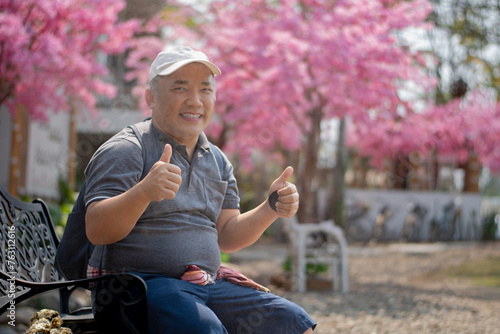 Asian middle-aged man in white cap and has backpack standing and thumping up in front of sakura flower tree, soft focus, happiness of middle-aged people and a single man around the world concept. photo