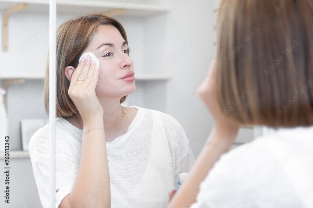 Attractive woman looking at mirror and applying moisturizing cream on cheeks in bathroom. Groomed young girl doing skincare morning routine after taking a shower Attractive woman looking at mirror and applying moisturizing cream on cheeks in bathroom. Groomed young girl doing skincare morning routine after taking a shower