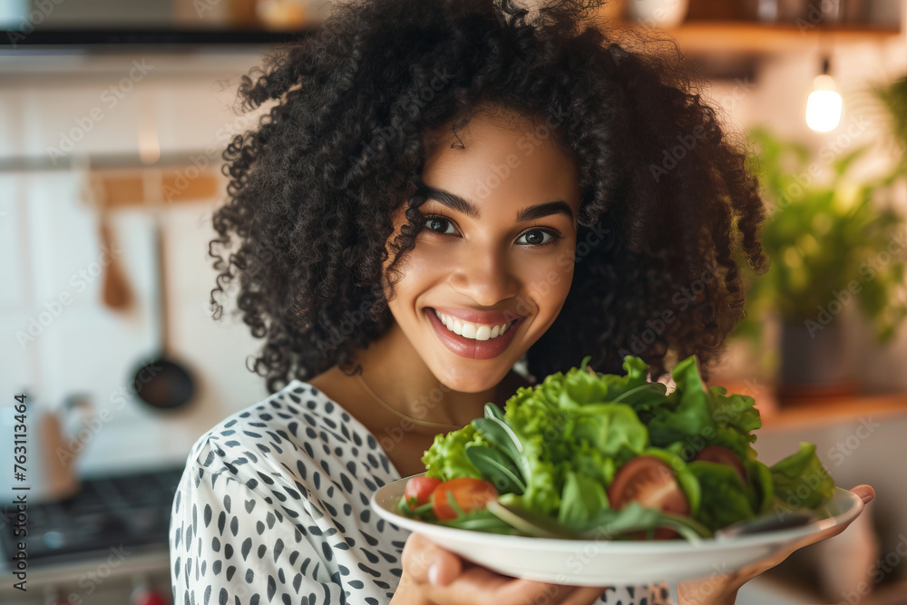 Cute, young girl in kitchen holding plate with salad of fresh vegetables, herbs and smiling
