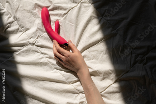 female hand reaching for a pink sex toy rabbit shaped vibrator for women in bed, sun rays on a quilted white blanket, good morning concept