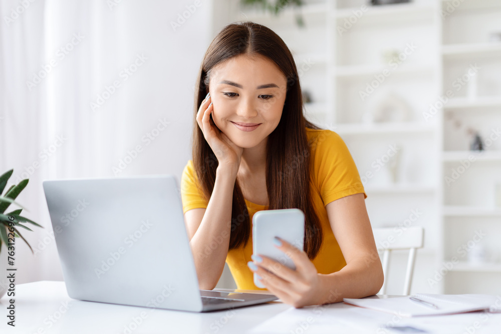Smiling asian woman looking at her smartphone while sitting in home office