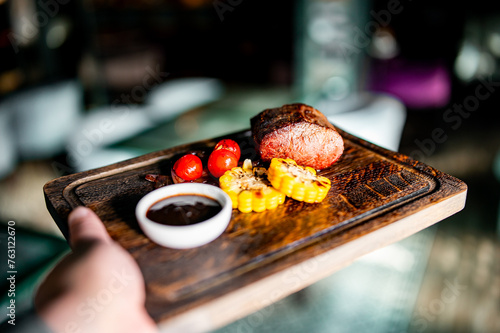 close-up of a hand holding a wooden serving board with food. A succulent piece of grilled steak is the centerpiece, cooked to perfection and showcasing an appetizing sear
