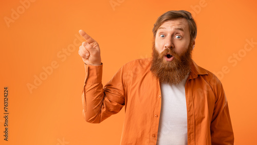 Amazed bearded man pointing at empty space over orange background