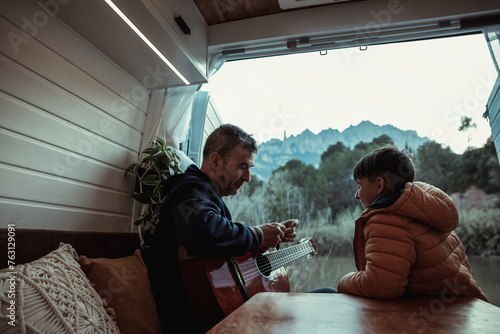  Father playing guitar for his son at sunset while sitting in a vintage Nordic style camper van. Conceptual van life, family, people © Laura