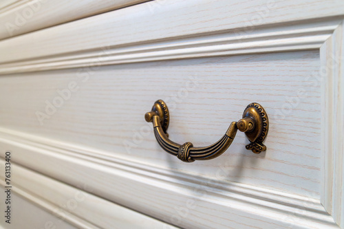 A bronze color handle on a white dresser. The handle is ornate