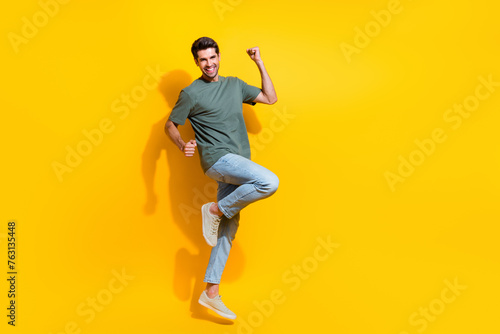 Full length photo of positive lucky man dressed khaki t-shirt rising fists jumping high empty space isolated yellow color background