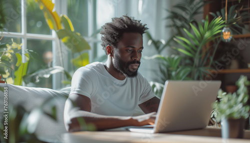 Close up of black american man in white tshirt sitting and typing, working on laptop keyboard in bright scandinavian interior of living room. Concept of freelance work at home, studying at home.