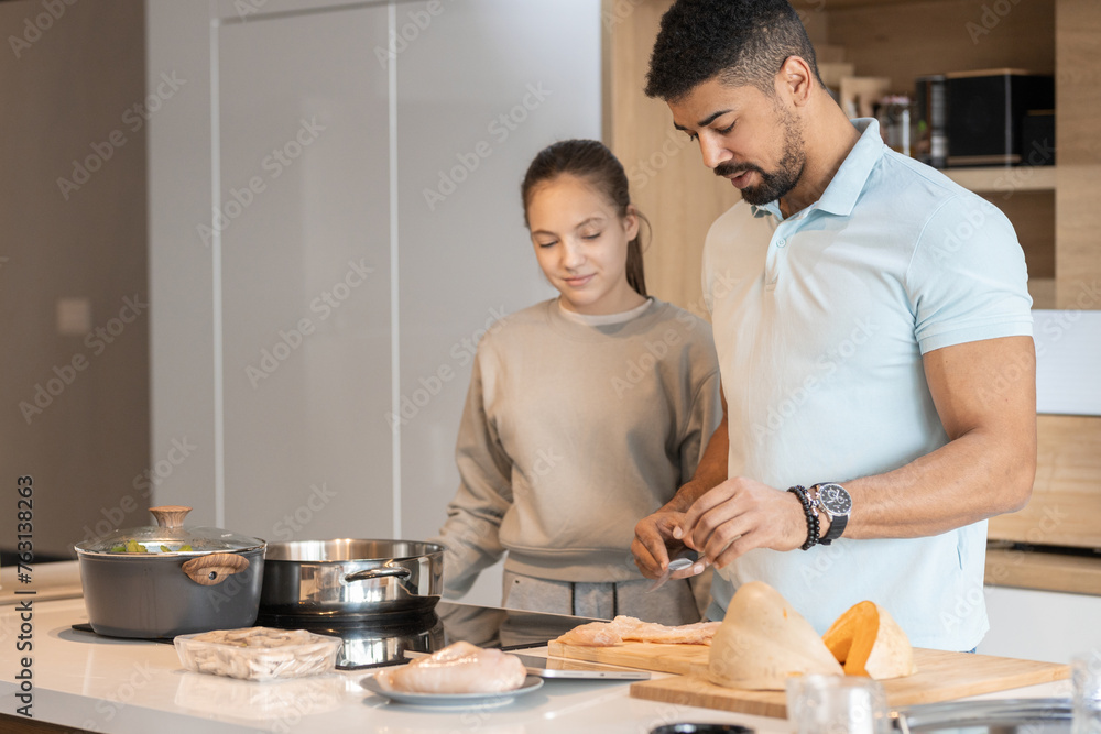 Daughter watching her dad while he cutting meat and preparing food in the kitchen at home Daughter watching her dad while he cutting meat and preparing food in the kitchen at home