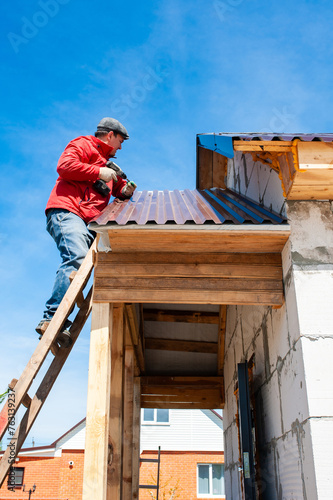 A worker builds a roof in a house while standing on a wooden ladder. Blue sky