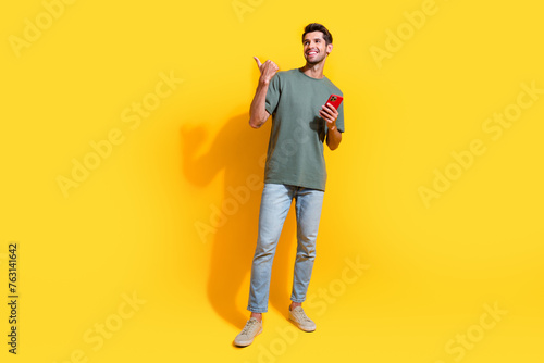 Full length photo of cheerful dreamy guy wear khaki t-shirt chatting modern device thumb empty space isolated yellow color background
