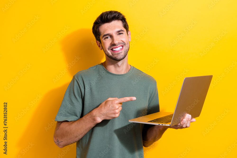 Photo of clever cheerful man with bristle dressed khaki t-shirt indicating at laptop in arm isolated on vibrant yellow color background Photo of clever cheerful man with bristle dressed khaki t-shirt indicating at laptop in arm isolated on vibrant yellow color background