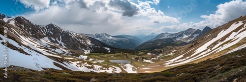 Panoramic view of a vast alpine landscape with lingering snow patches, a small pond, and mountain ridges under a partly cloudy sky © mikeosphoto