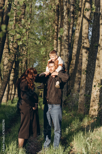 A husband walks in the park with his pregnant wife and holds their son on his shoulders, enjoying family time together. © Nastassia