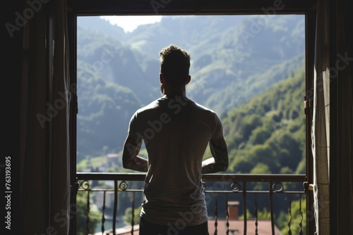 Backlit silhouette of a man in a simple t-shirt, standing before a window with a panoramic view of a mountainous landscape. The silhouette reveals the contours of his tattooed arms.