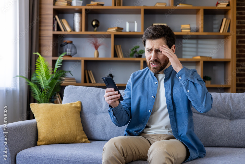 Worried man sitting on couch looking at smartphone Worried man sitting on couch looking at smartphone
