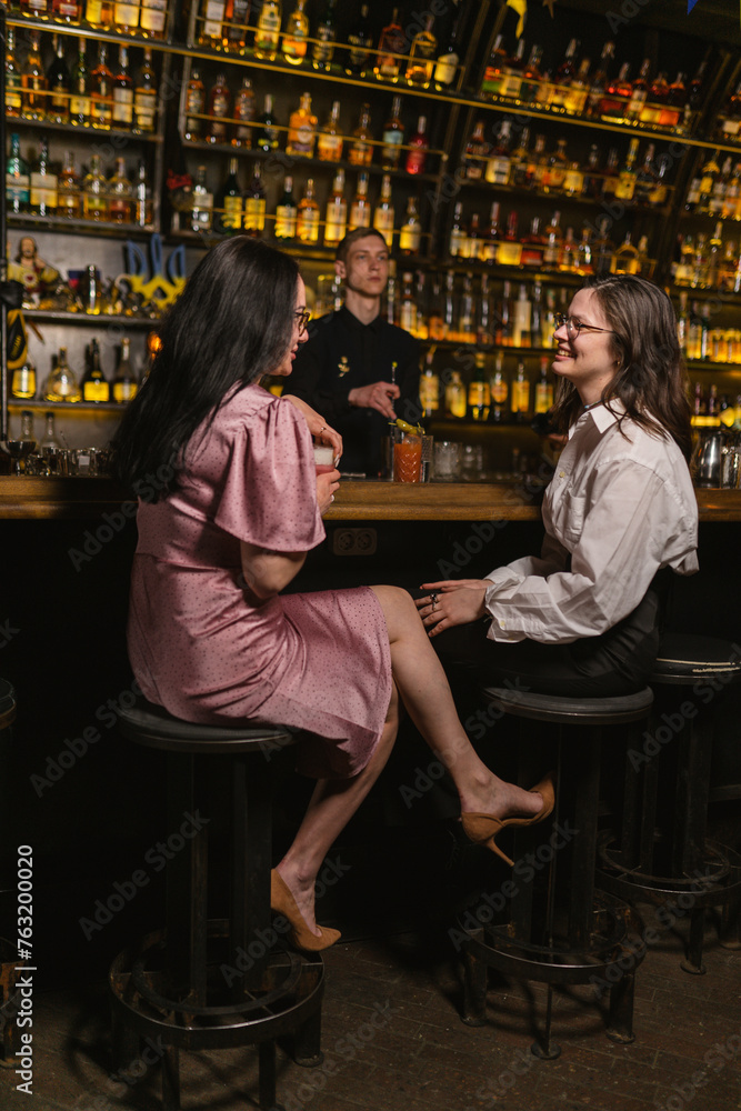 Young ladies flirt with bartender at bar counter. Pleasant meeting in fancy place with various bottles of alcohol in club