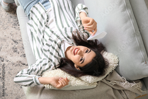 Happy young woman with paper plane lying on sofa at home