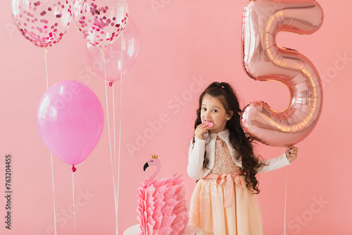 Adorable little girl in a pink dress posing next to a flamingo birthday cake and celebrating her fifth birthday on a pink background photo