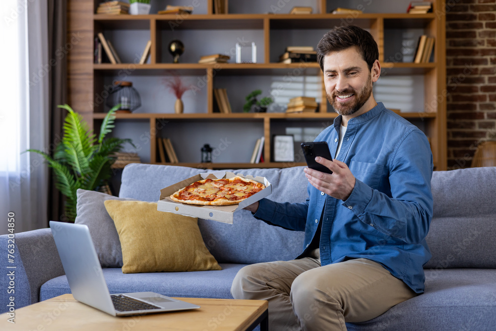 Smiling man enjoying pizza while using smartphone at home Smiling man enjoying pizza while using smartphone at home