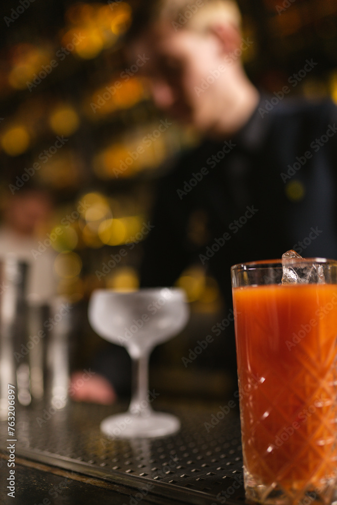 Cocktail with ice cubes on bar counter with glass on blurred background of bartender in club. Concept of finished drink for visitor and specialist workflow