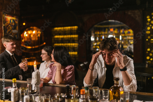 Drunk guy feels unwell after cocktail party at bar. Man sits apart from friends and holds head near bar counter at night