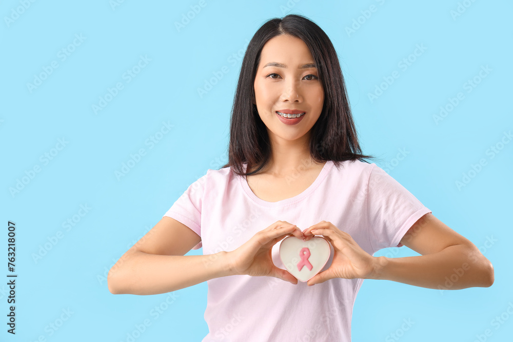 Young Asian woman with cookie making heart gesture on blue background. Breast cancer awareness concept Young Asian woman with cookie making heart gesture on blue background. Breast cancer awareness concept