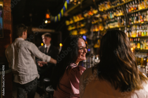 Happy brunette lady with friend rest together in alcohol bar. Young women enjoy meeting at party against shelves with bottles