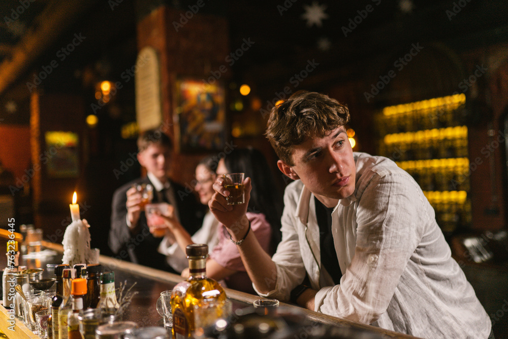 Tired drunk man with shot glass sits at wooden bar counter. Guest suffers from alcoholism spending time in popular night club - obrazy, fototapety, plakaty 