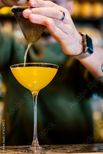 bartender meticulously pouring a bright yellow cocktail through a metal strainer into a martini glass. The scene exudes professionalism and craftsmanship, with attention to detail in presentation