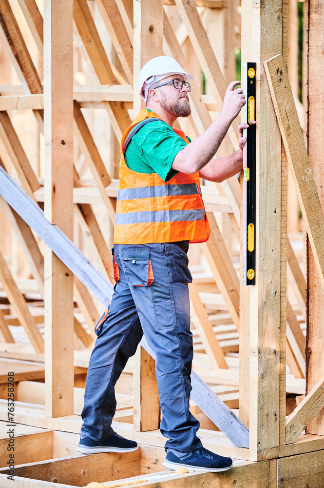 Carpenter constructing two-storey wooden frame house. Man inspects walls for levelness using spirit level, wearing protective overalls, helmet and vest. Concept of modern ecological construction. Carpenter constructing two-storey wooden frame house. Man inspects walls for levelness using spirit level, wearing protective overalls, helmet and vest. Concept of modern ecological construction.