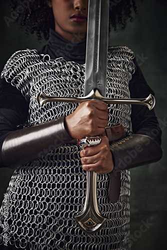 Close-up of African woman in chainmail holding sword vertically in front of face against vintage green background. Focus on hands and sword. Concept of history, beauty and fashion, comparison of eras