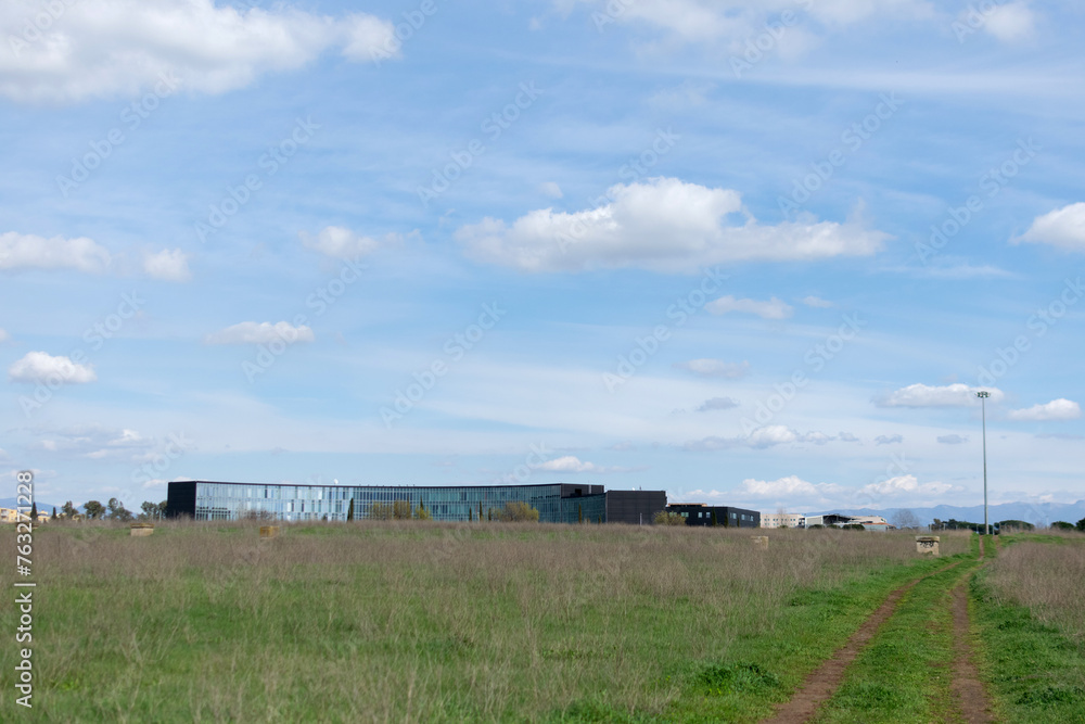 Horizontal landscape - under the blue sky on the horizon the mirrored long modern building, seen across a field with green grass