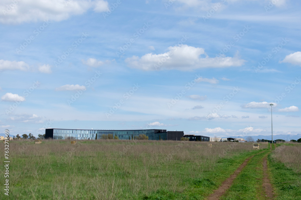 Horizontal landscape - under the blue sky on the horizon the mirrored long modern building, seen across a field with green grass
